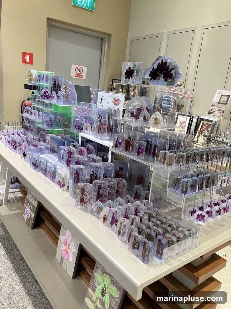A display of various items featuring preserved flowers, likely orchids, encased in clear resin blocks of different shapes and sizes. The items are arranged on tiered shelves. Some items are small cubes, while others are larger, irregular shapes. In the foreground, there are also framed prints of flowers. In the background, there is a doorway with an 'EXIT' sign and a 'No entry' sign.