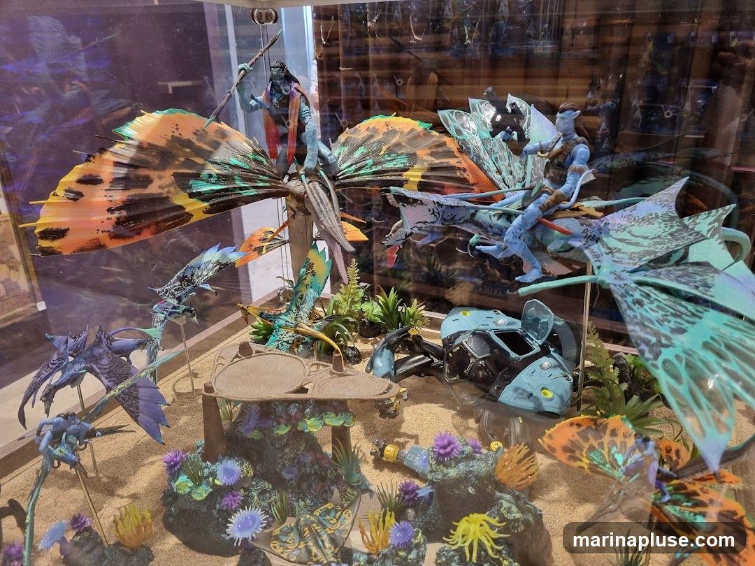 A diorama featuring figures and creatures from the movie Avatar, displayed in a glass case. The scene includes Na'vi figures riding winged creatures, smaller flying creatures, and a crashed vehicle. The diorama is set on a sandy base with colorful coral and plant life.