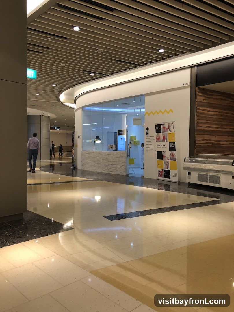 A view of a medical clinic's storefront in a modern indoor shopping area. The clinic has a glass facade, allowing a view into the reception area with a desk and a lamp. A poster on the wall displays information about "PAIN INJURY MANAGEMENT" and "CHINESE MEDICINE". The floor is polished with a reflective surface, and other shops and walkways are visible in the background.