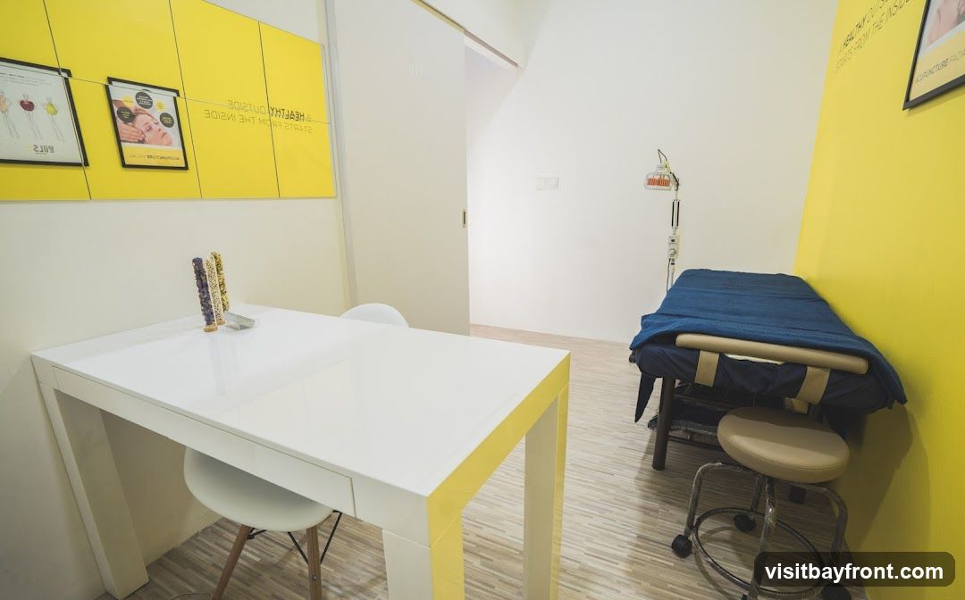 A bright and clean treatment room with yellow accent walls. A white table with decorative items sits in the foreground, with two chairs. In the background, a massage table covered with a blue blanket is positioned next to a heat lamp. A poster with text and images is visible on the wall.