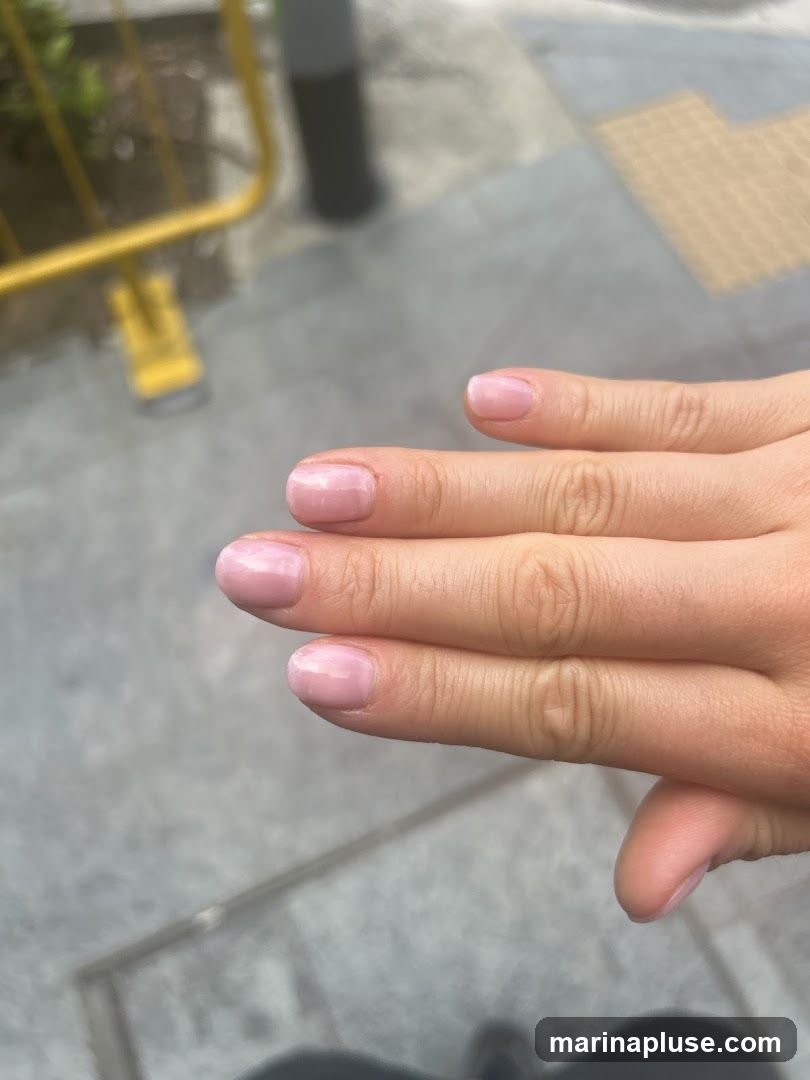 A close-up shot of a person's hand with light pink, glossy nails. The background is blurred and appears to be an outdoor setting with a grey paved ground and a yellow barrier.