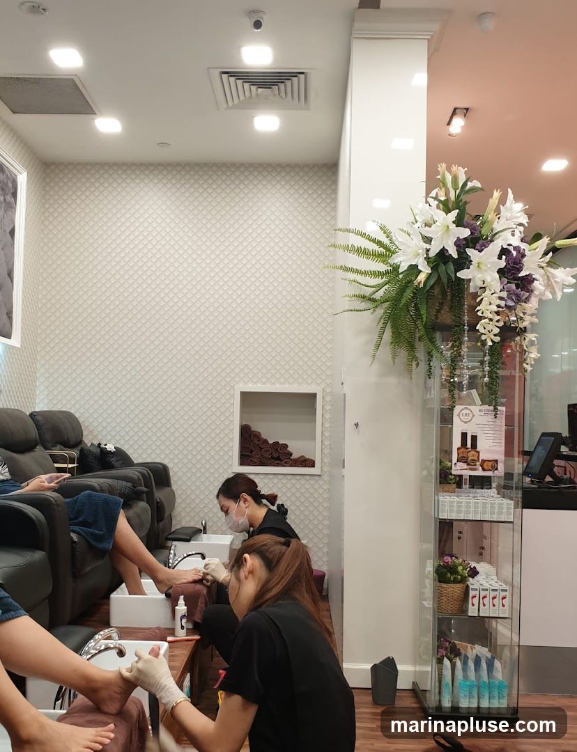 A nail salon interior with several clients receiving pedicures. Two technicians are working on clients' feet, while another client relaxes in a recliner. A display shelf holds various nail care products and a floral arrangement adorns a pillar.