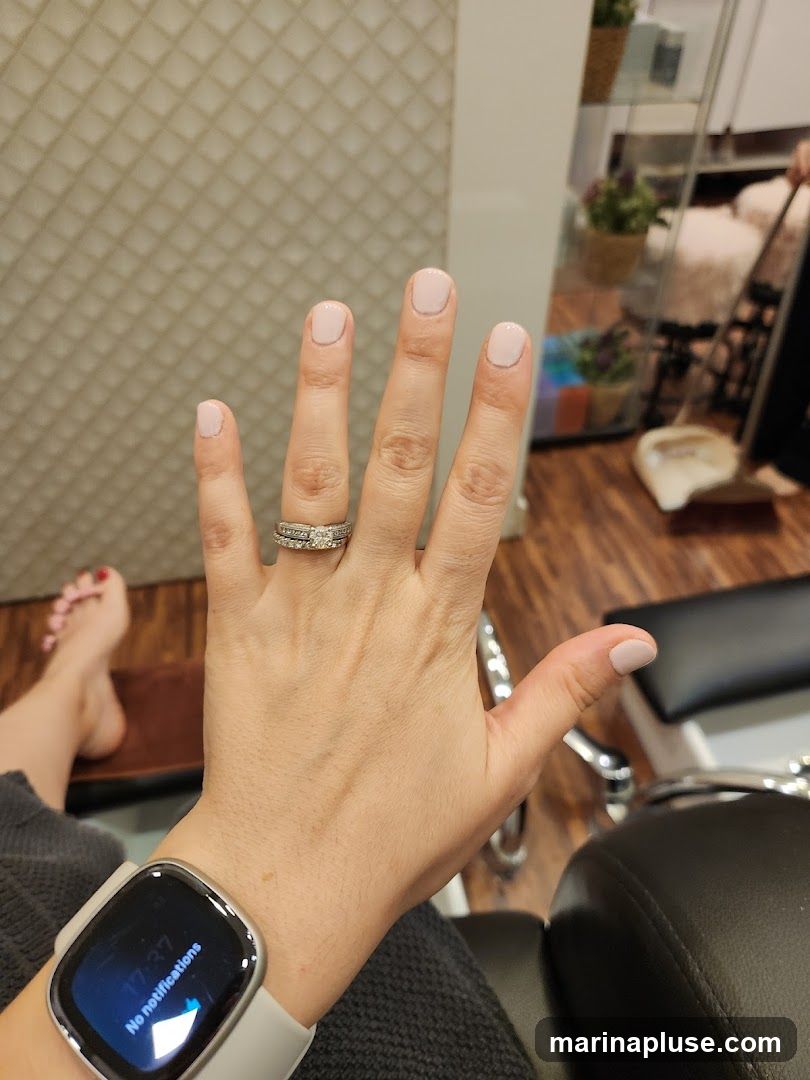 A person's hand with a light pink manicure is shown, with a smartwatch on the wrist. In the background, a pedicure chair and a shelf with decorative plants are visible, suggesting a nail salon environment.
