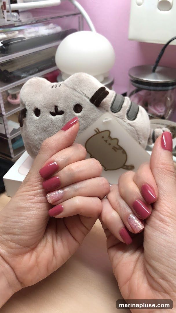 A close-up shot of a person's hands with a manicure. The nails are painted in a deep pinkish-red color, with accent nails on the ring fingers featuring a glittery pink polish. In the background, a plush toy resembling the character Pusheen the cat is visible, along with a desk lamp and organizers.