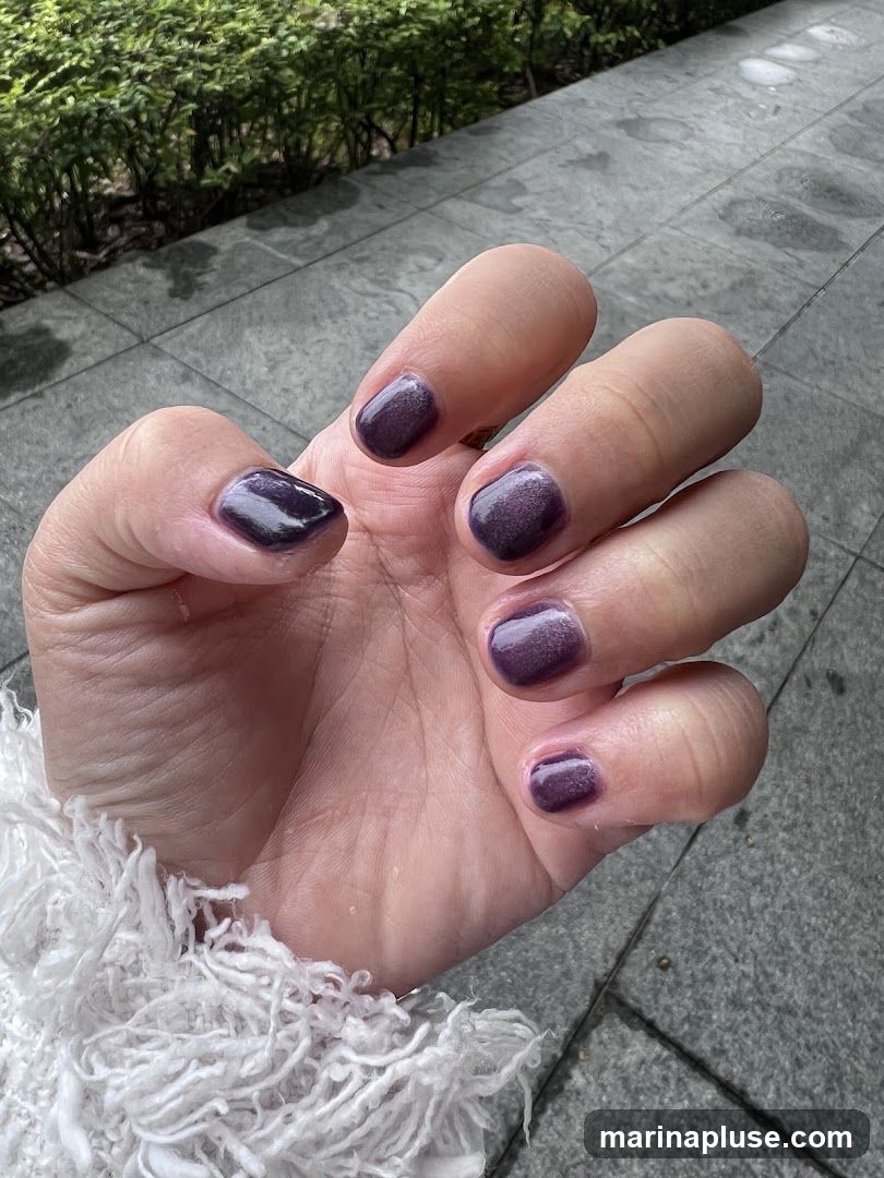 A close-up shot of a woman's hand with short, dark purple, shimmery nail polish. The hand is resting on a textured white garment. The background shows a tiled walkway and green foliage.