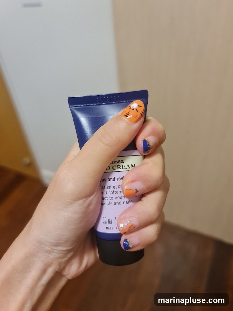 A hand with decorative nail art is holding a tube of hand cream. The nail art includes orange and blue colors, with some glitter and a tiger face design on one nail. The hand cream tube is purple and has text on it, including "Aissa HAND CREAM" and "30 ml".