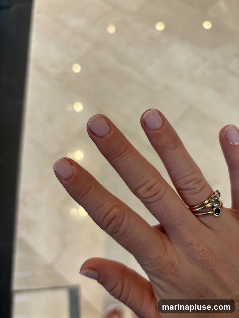 A close-up shot of a person's hand with light pink, freshly manicured nails. The hand is adorned with several stacked gold rings, one of which features small dark and light stones. The background appears to be a polished, light-colored floor with soft, out-of-focus circular reflections.