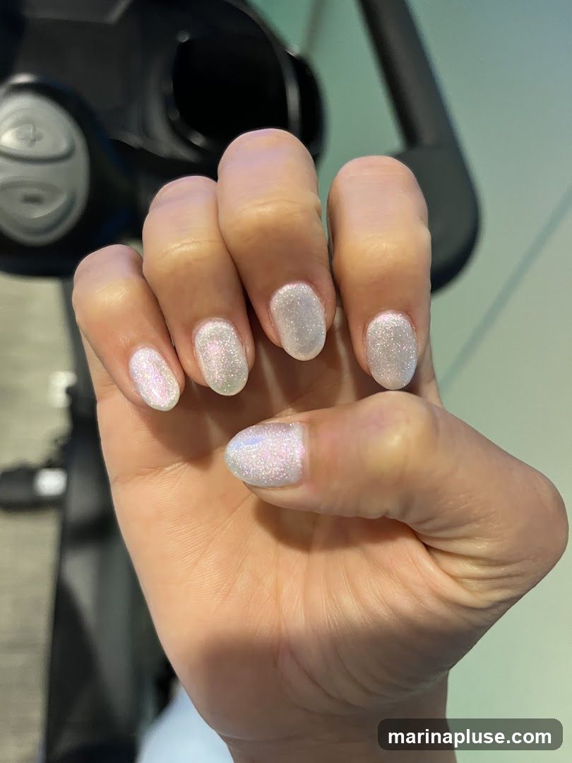 A close-up shot of a person's hand with short, oval-shaped nails. The nails are painted with a shimmering, iridescent white polish that catches the light, giving them a subtle holographic effect. The background is blurred, showing a dark, curved object and a light green wall.