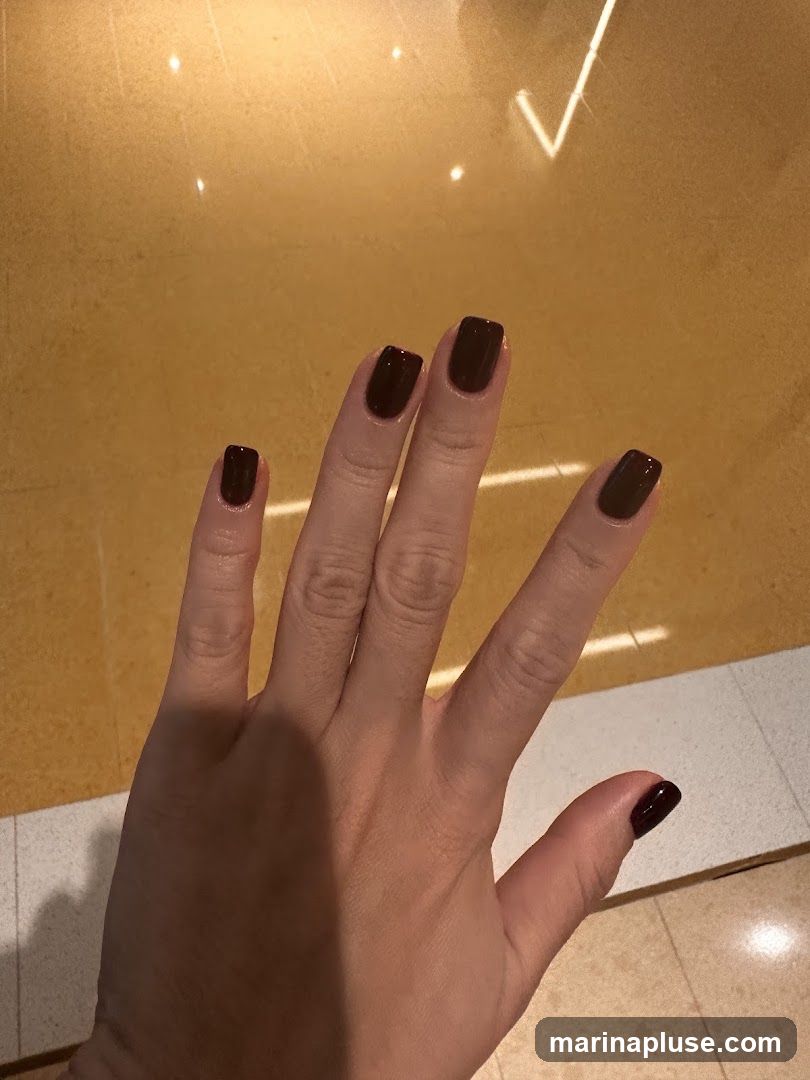 A hand with dark red, square-shaped fingernails is shown against a yellow, reflective surface. The lighting creates bright reflections on the surface.