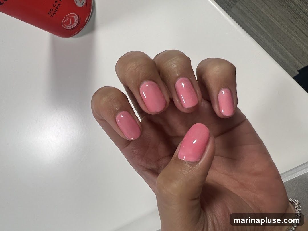 A close-up shot of a person's hand with neatly manicured nails painted in a bright pink shade. The nails are oval-shaped and have a glossy finish. A portion of a red object with some text and a circular logo is visible in the upper left corner.
