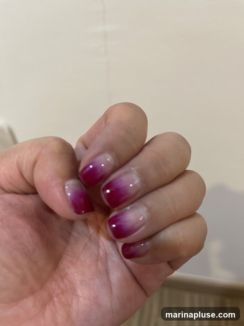 A close-up shot of a hand with ombre-style fingernails. The nails transition from a clear or light pink at the base to a deep magenta or fuchsia at the tips. The background is a plain, light-colored wall.