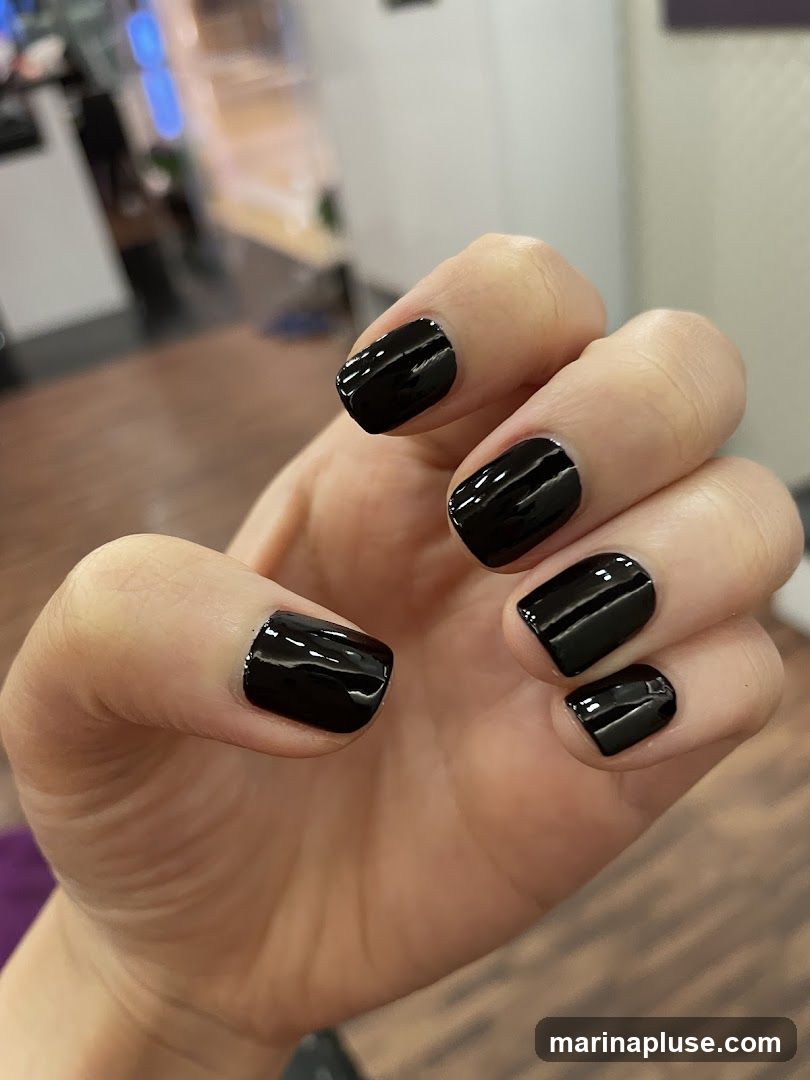 A close-up shot of a hand with a fresh black manicure. The nails are short and square-shaped, with a high-gloss finish. The background is blurred, showing a wooden floor and what appears to be a salon interior.