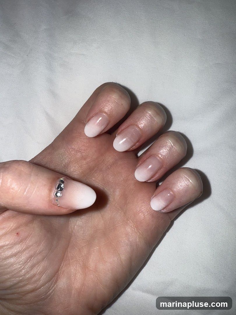 A close-up shot of a hand with manicured nails. The nails feature an ombre design, transitioning from a sheer nude base to a white tip. The ring finger nail is adorned with a line of small, clear rhinestones. The background is a white, textured fabric.