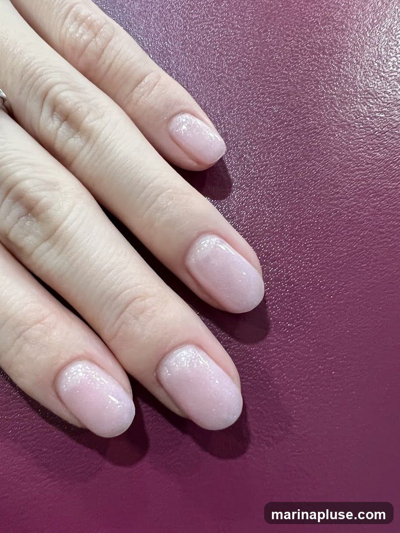 A close-up shot of a person's hands with light pink, glitter-infused gel nails. The nails have a subtle shimmer and are neatly shaped. The background is a textured, deep purple surface.