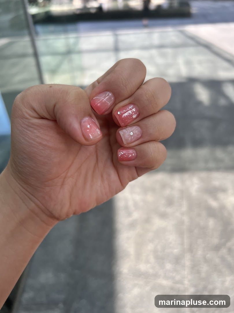 A close-up of a hand with short, square-shaped nails. The nails are decorated with a gradient of pink and white polish, with some nails featuring glitter accents and small floral designs. One nail has a tiny rhinestone embellishment. The background is blurred and appears to be an outdoor setting with sunlight casting shadows.