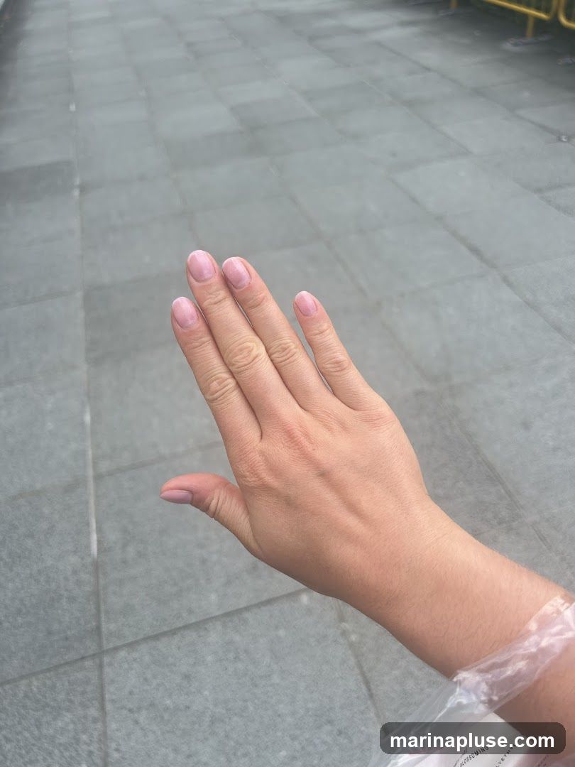 A person's hand with light pink, natural-looking nails is held up against a background of grey tiled pavement. The hand is positioned in the center of the frame, with the fingers slightly spread. The skin appears smooth and unblemished. A clear plastic bag is visible around the wrist, partially obscuring the lower arm. The pavement tiles are square and laid in a grid pattern. A yellow barrier is faintly visible in the upper right corner of the image.