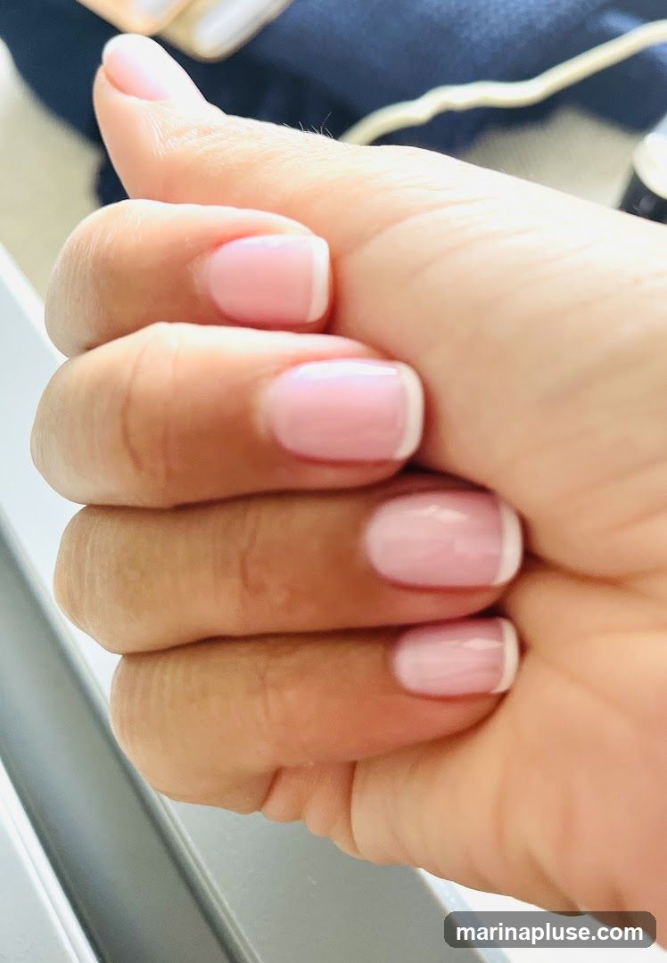 A close-up shot of a hand with a fresh French manicure. The nails are neatly shaped and painted with a sheer pink base and a crisp white tip. The cuticle area appears clean and well-maintained. The background is blurred, with hints of dark fabric and a light-colored surface.