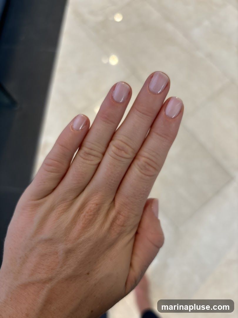 A close-up shot of a person's hand with neatly manicured nails. The nails are painted with a sheer, pale pink or nude polish, giving them a natural and clean appearance. The hand is positioned against a blurred background of a light-colored, possibly tiled floor.