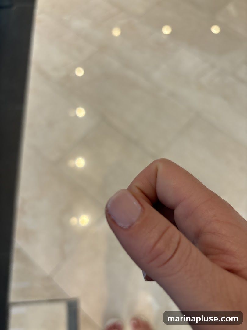 A close-up shot focuses on a person's hand, with their thumb and index finger held close together. The background is blurred, showing a light-colored tiled floor with reflections of lights. The person is wearing light-colored shoes, visible at the bottom of the frame.