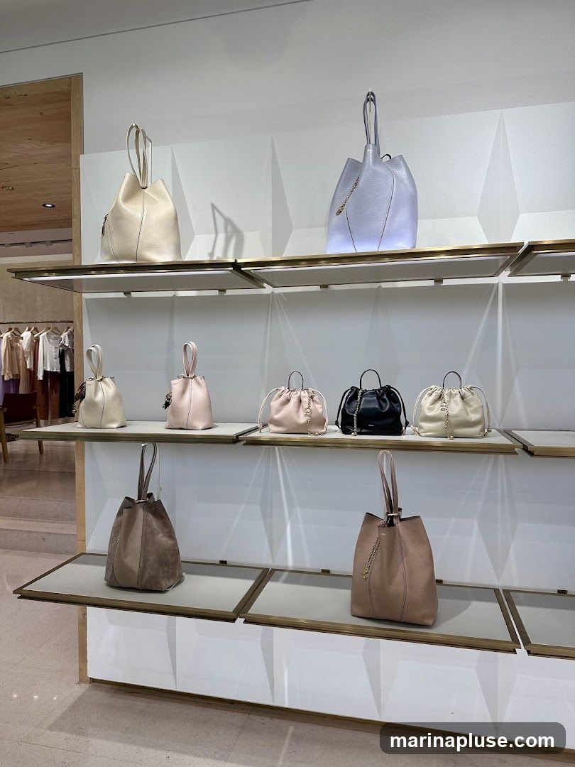 A retail display featuring several handbags on tiered shelves. The bags are in various neutral colors including beige, light pink, black, and a light purple-grey. Some bags have chain detailing. In the background, clothing racks with garments are visible.