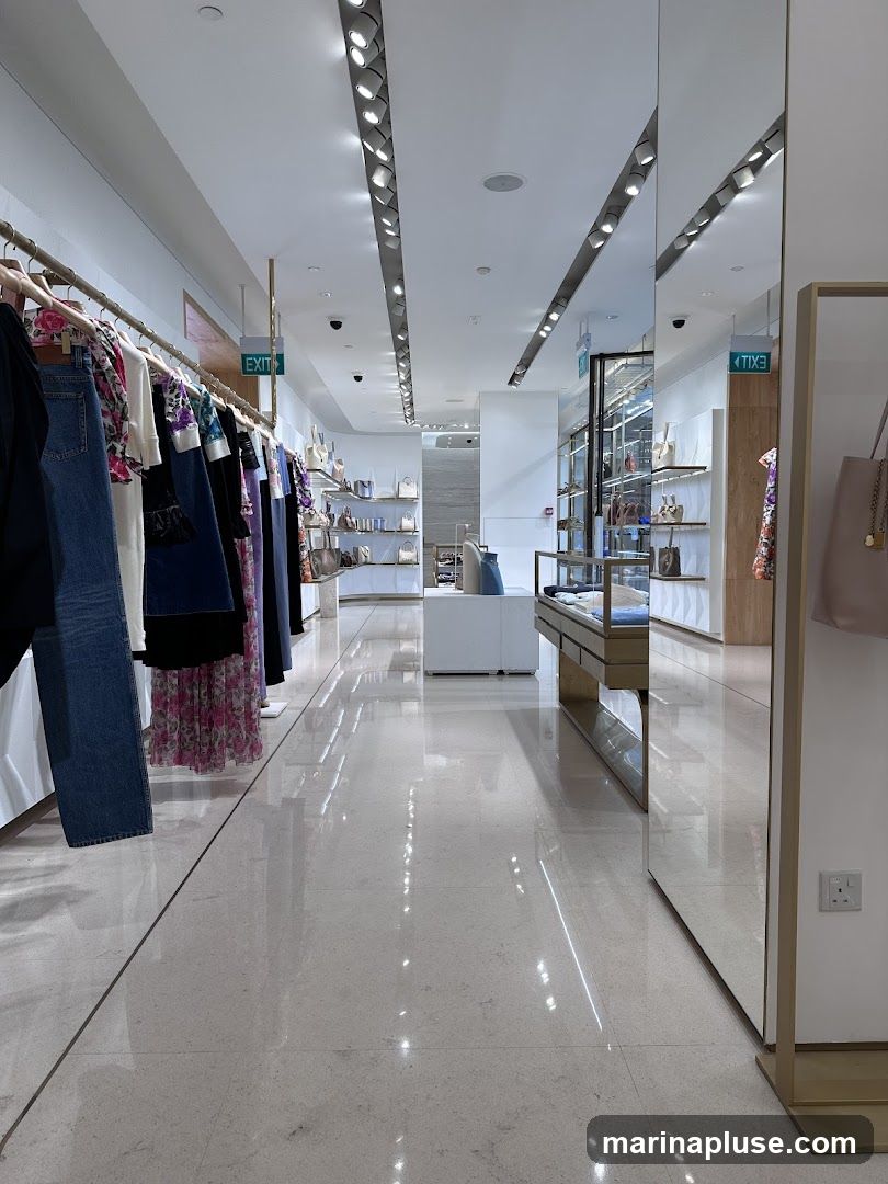 A well-lit, modern clothing store interior with racks of apparel and display cases. The floor is highly polished, reflecting the ceiling lights and merchandise. On the left, clothing items hang on a rack, including jeans, patterned tops, and dresses. Further down the aisle, shelves display handbags and accessories. A glass display case showcases more items. An 'EXIT' sign is visible on the ceiling.