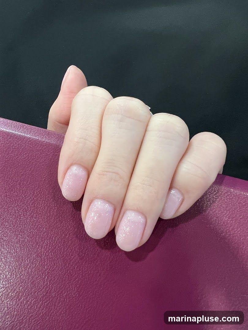 A close-up shot of a person's hand with neatly manicured nails. The nails are painted in a light, sheer pink polish with a subtle glitter or shimmer effect. The hand is resting on a textured, deep purple surface, possibly a book cover or a piece of fabric. The background is a solid black.