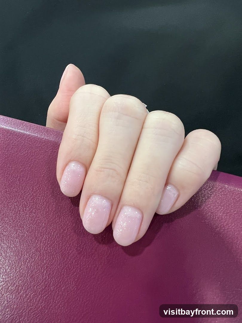 A close-up shot of a person's hand with neatly manicured nails. The nails are painted in a light, sheer pink polish with a subtle glitter or shimmer effect. The hand is resting on a textured, deep purple surface, possibly a book cover or a piece of fabric. The background is a solid black.