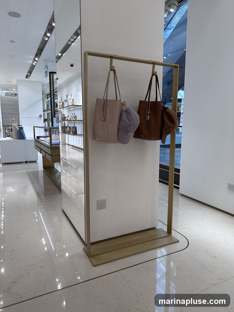 A retail display featuring two handbags and two hats. One handbag is light pink and the other is brown. Both are hanging on a gold-colored rack. A light purple cap is hanging from the pink handbag, and a brown cap is hanging from the brown handbag. The display is set against a white panel with a mirrored surface to the left, reflecting the interior of a store. The floor is tiled with a light-colored terrazzo pattern.