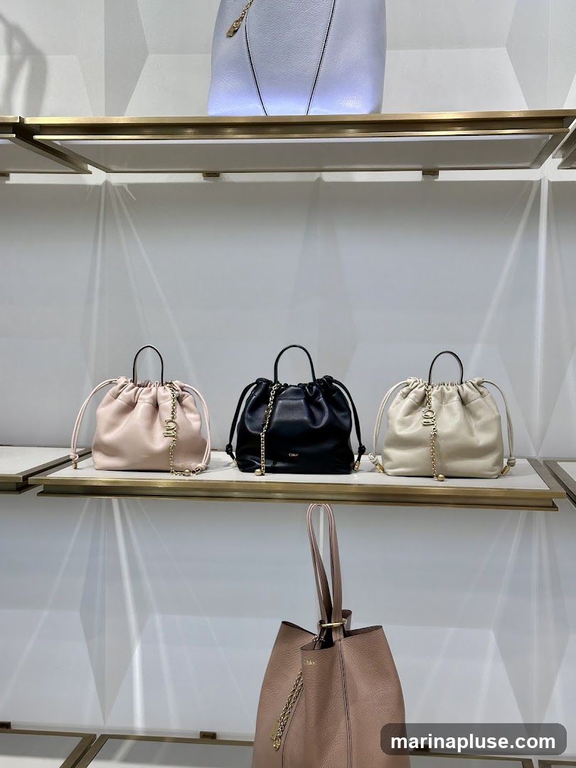 A display of several handbags on white shelves. The top shelf shows a light blue handbag. The middle shelf displays three smaller drawstring handbags in pale pink, black, and cream. The bottom shelf features a larger pale pink tote bag. All handbags appear to be designer items, with visible branding on some.