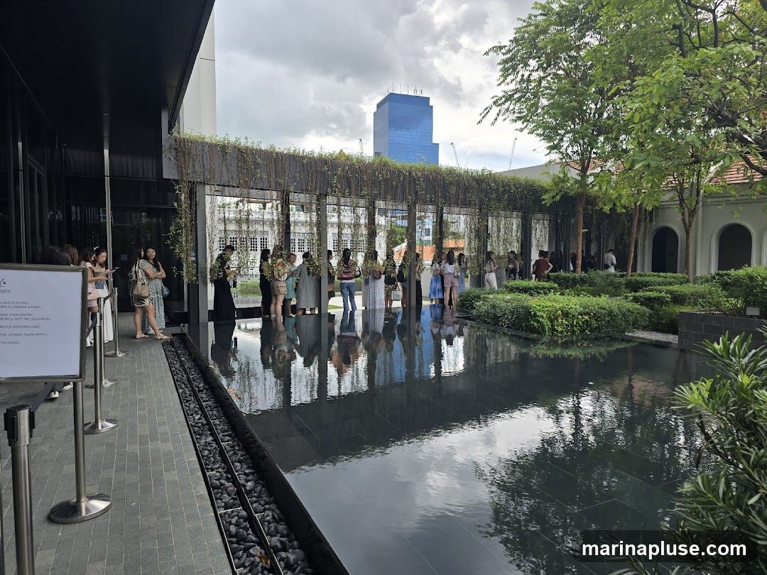 A group of people are gathered outdoors near a building with a reflective pool in the foreground. The pool mirrors the surrounding greenery and the sky. A sign is visible on the left, and lush plants and trees are scattered throughout the scene. In the background, a modern skyscraper stands against a cloudy sky.