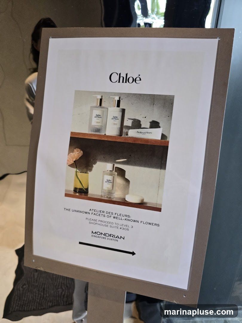 A sign displays an advertisement for Chloé's Atelier Des Fleurs collection, featuring perfume bottles and a tube of hand cream on shelves. Text below the image indicates that the collection is called 'The Unknown Facets of Well-Known Flowers' and directs visitors to proceed to Level 3, Shophouse Suite #305 at Mondrian Singapore Duxton. An arrow points to the right.