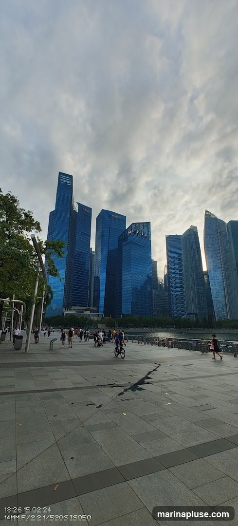 A view of a modern cityscape with tall, blue-glass skyscrapers under a cloudy sky. In the foreground, a wide paved promenade runs along a waterfront, with several people walking and cycling. Trees line the left side of the promenade. The scene appears to be taken during the late afternoon or early evening.