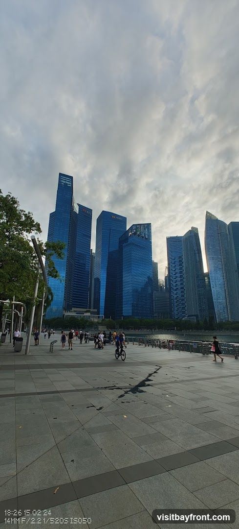 A view of a modern cityscape with tall, blue-glass skyscrapers under a cloudy sky. In the foreground, a wide paved promenade runs along a waterfront, with several people walking and cycling. Trees line the left side of the promenade. The scene appears to be taken during the late afternoon or early evening.