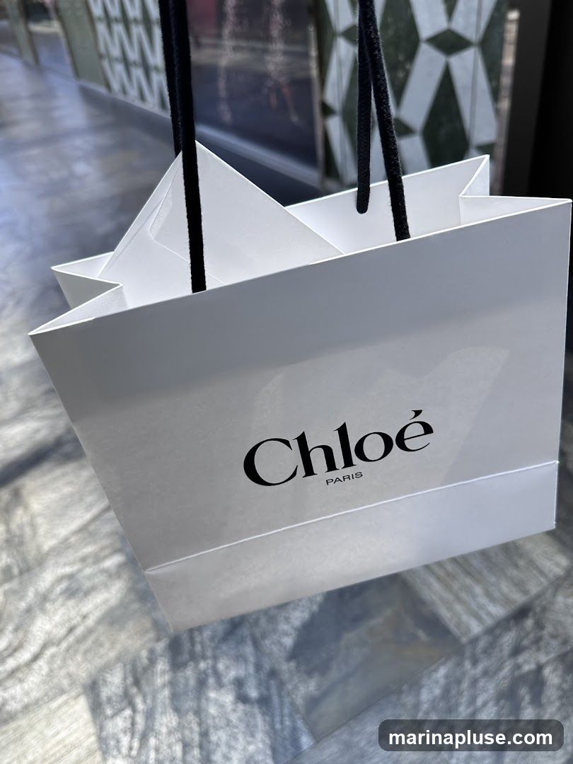 A white Chloe shopping bag with black handles and the Chloe Paris logo printed on the front. The bag appears to be full, with white tissue paper peeking out from the top. The background shows a blurred interior with patterned walls and a tiled floor.
