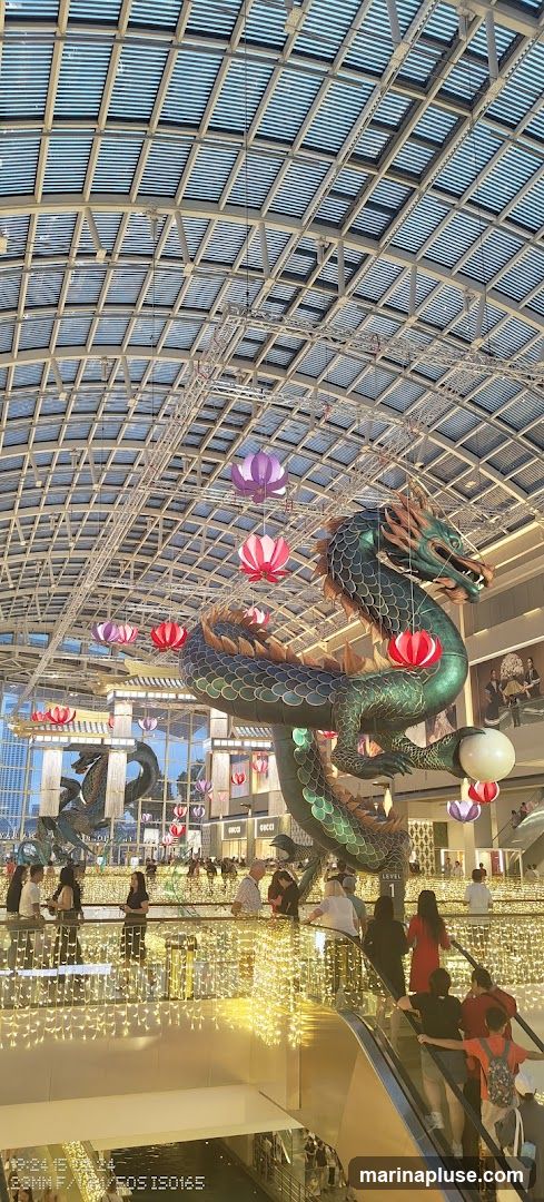 A large, green dragon sculpture hangs from the ceiling of a mall, adorned with colorful paper lanterns. Below, people are seen on escalators and walkways, surrounded by strings of warm, yellow lights. The mall features a modern, open-air design with a glass roof and visible structural beams. Storefronts, including one with a visible 'GUCCI' sign, are present in the background.