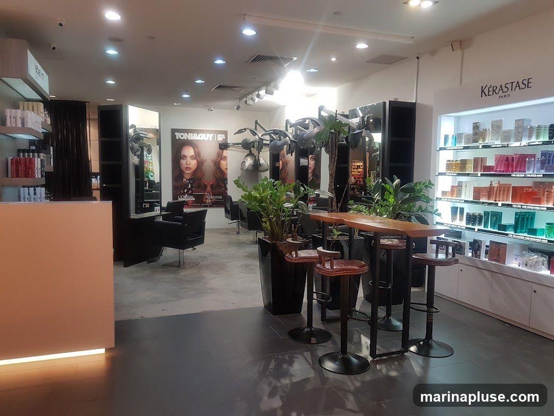 The interior of a hair salon features a reception desk, styling stations with chairs and hair dryers, and shelves stocked with hair products. A high table with stools is set up in the foreground, and a large poster for Toni & Guy is visible on the wall. The Kérastase Paris brand is prominently displayed on a shelf unit filled with various hair care products.