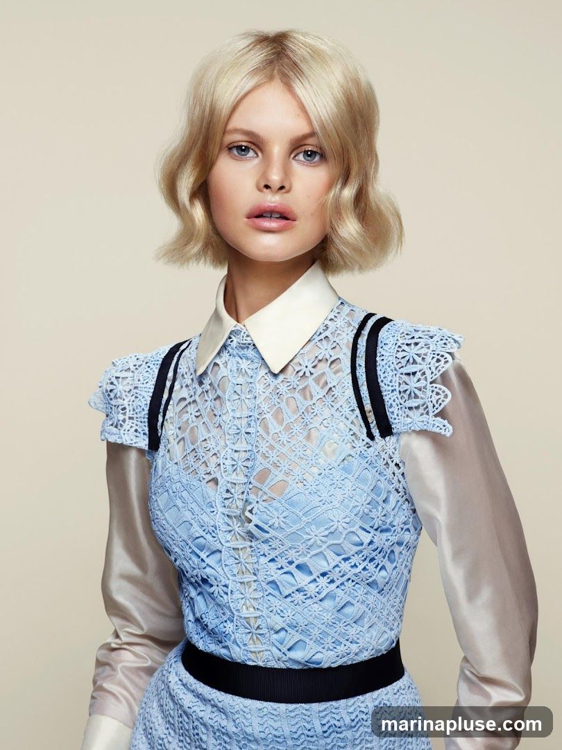 A fashion model with blonde wavy hair and blue eyes poses against a neutral background. She is wearing a light blue lace dress with a white collar and sheer, light-colored sleeves. The dress features intricate detailing and black ribbon accents.