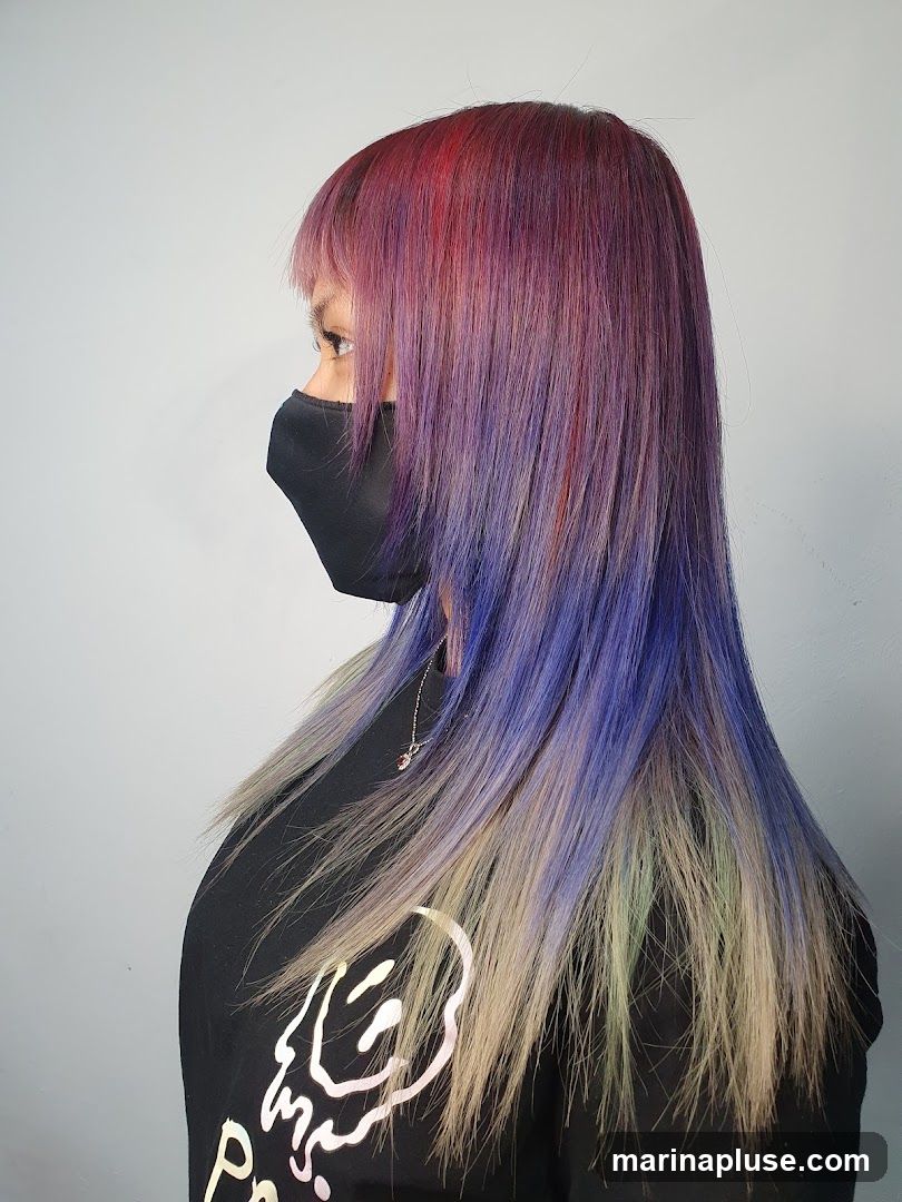 A woman with long, layered hair dyed in shades of red, purple, blue, and blonde. She is wearing a black face mask and a black t-shirt with a graphic print. The background is a plain gray wall.