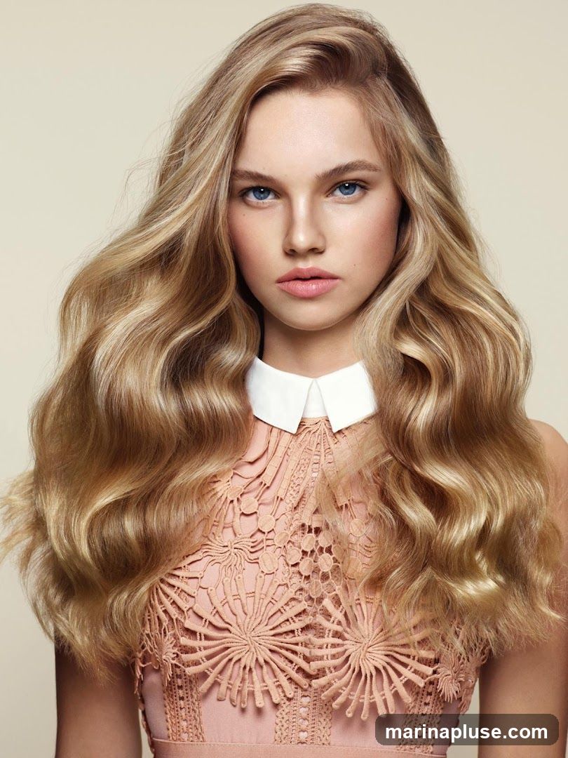 A close-up studio portrait of a young woman with long, wavy blonde hair. She has blue eyes and is wearing a peach-colored dress with a white collar. The background is a plain, light beige.