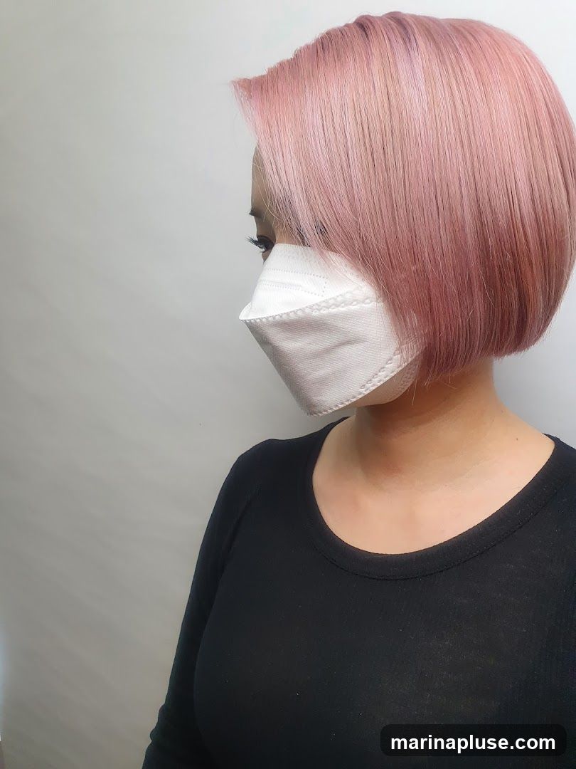 A woman with a short, pink bob haircut and a white mask is shown in profile against a plain background. She is wearing a black long-sleeved shirt.