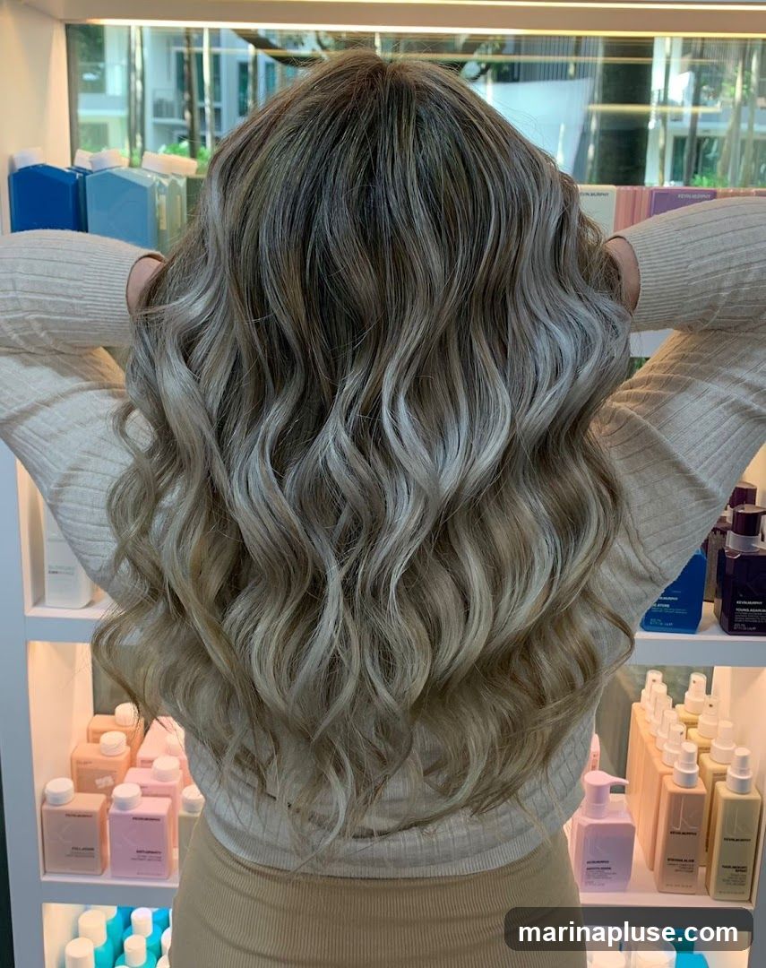 A woman is shown from behind, showcasing her long, wavy blonde balayage hair. She is wearing a cream-colored sweater and a beige skirt. Behind her, shelves are stocked with various hair care products in bottles and containers of different colors, including pink, blue, and purple. Some bottles have visible branding, such as "KEVIN MURPHY". The setting appears to be a salon or a retail space selling hair products.