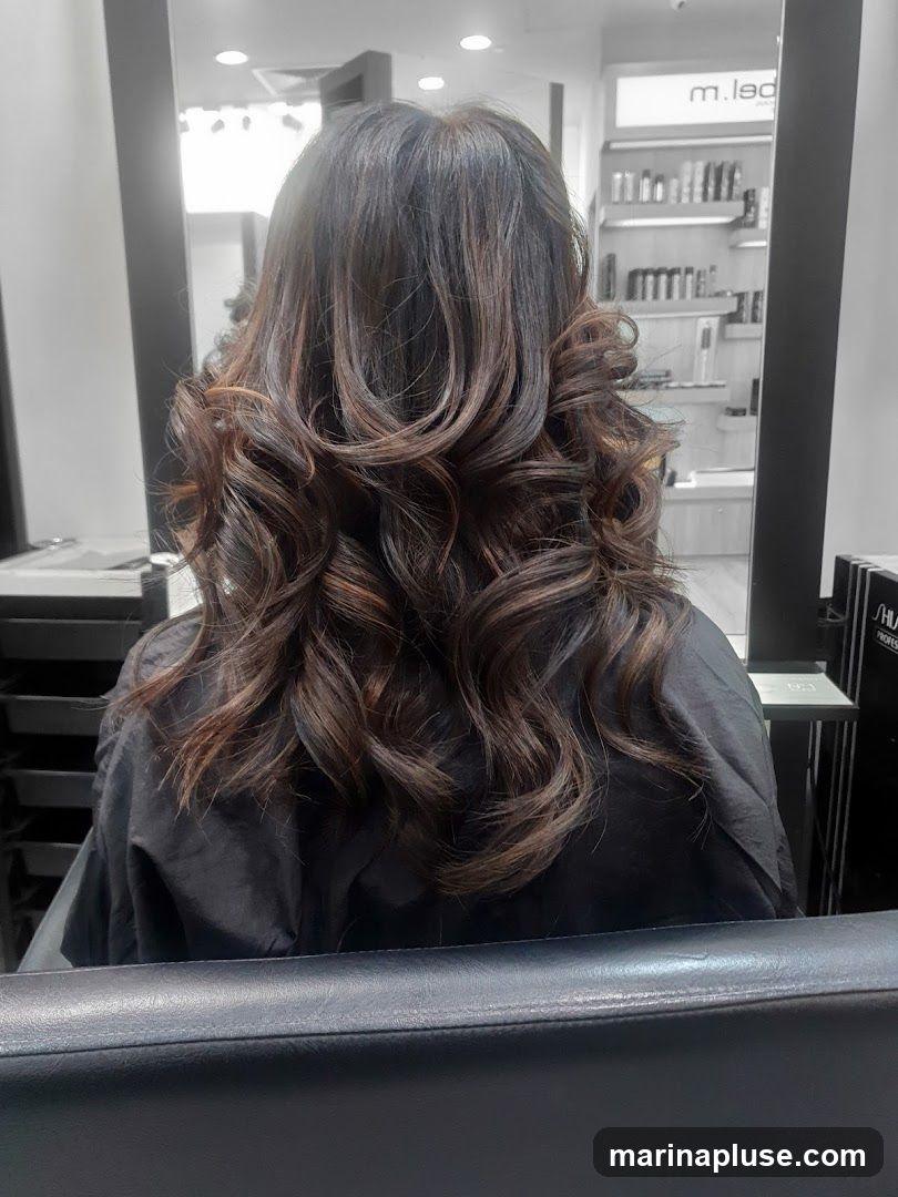 A woman's back is shown, with her long, dark hair styled in loose curls and highlights. She is wearing a black cape. In the background, shelves with hair products are visible, along with a sign that reads "m.led".