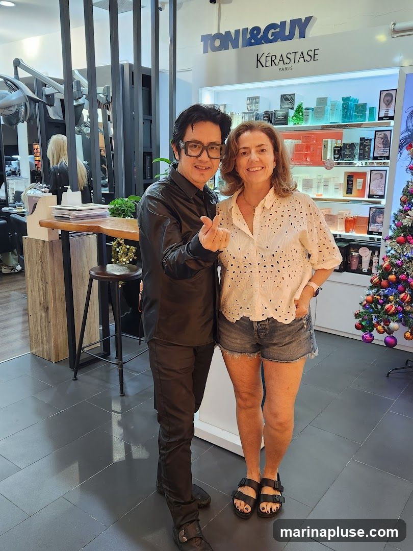 A man and a woman are posing for a photo inside a hair salon. The man, who appears to be a stylist, is wearing a black shirt and pants and glasses. He is making a heart shape with his fingers. The woman is wearing a white eyelet shirt and denim shorts. Behind them, shelves are stocked with hair products, and a Christmas tree is visible on the right. The salon's branding, "TONI&GUY KÉRASTASE PARIS," is displayed on the wall.