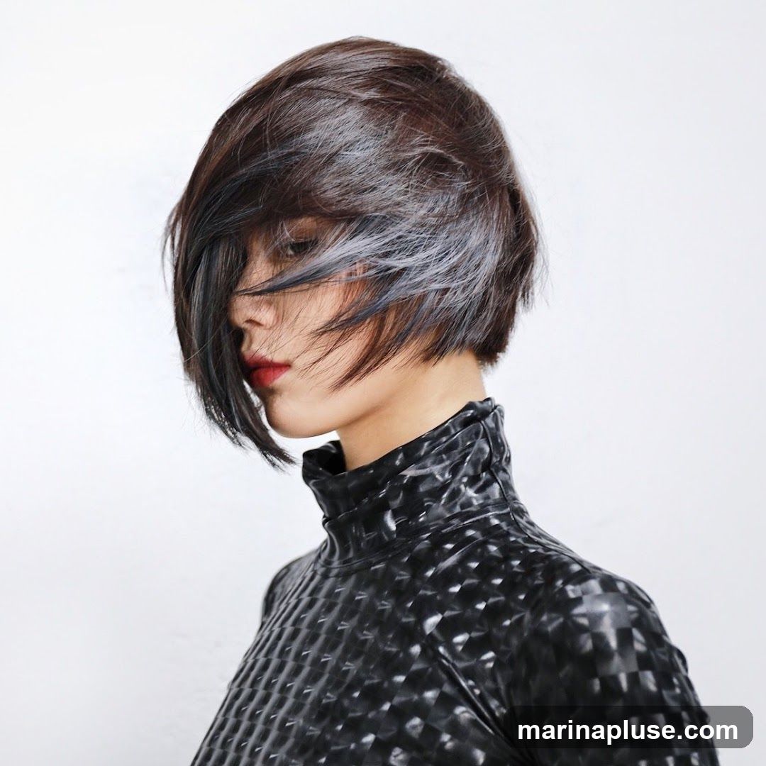 A profile view of a person with a short, dark, layered bob haircut. Streaks of grey are visible throughout the hair, and some strands fall across the face, partially obscuring the eyes. The person is wearing a black, high-necked garment with a textured, geometric pattern. Red lipstick is visible on the lips. The background is plain white.
