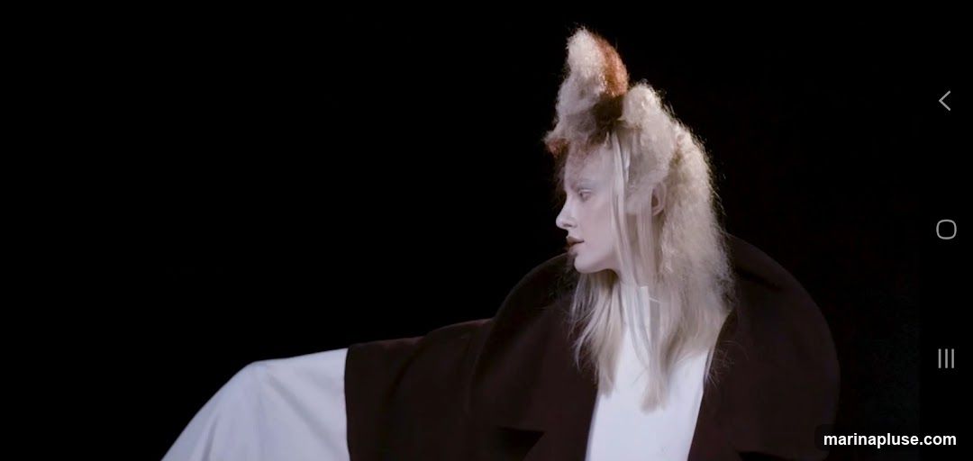 A model with pale skin and dramatic, avant-garde hair styled into a tall, textured shape with brown and blonde tones. They are wearing a dark coat over a white garment, against a black background.