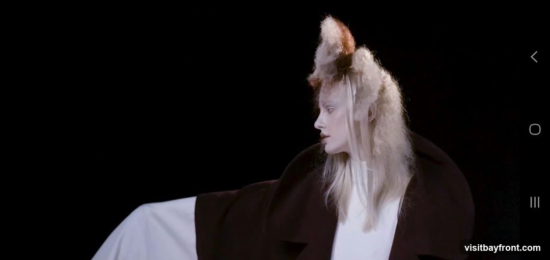 A model with pale skin and dramatic, avant-garde hair styled into a tall, textured shape with brown and blonde tones. They are wearing a dark coat over a white garment, against a black background.