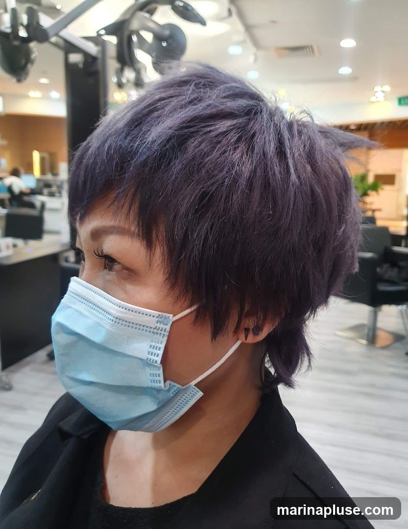 A woman with short, choppy purple hair and a light blue surgical face mask is shown in profile. She is wearing a black jacket and has small, dark triangular earrings. The background appears to be a salon or beauty parlor, with styling chairs and equipment visible.