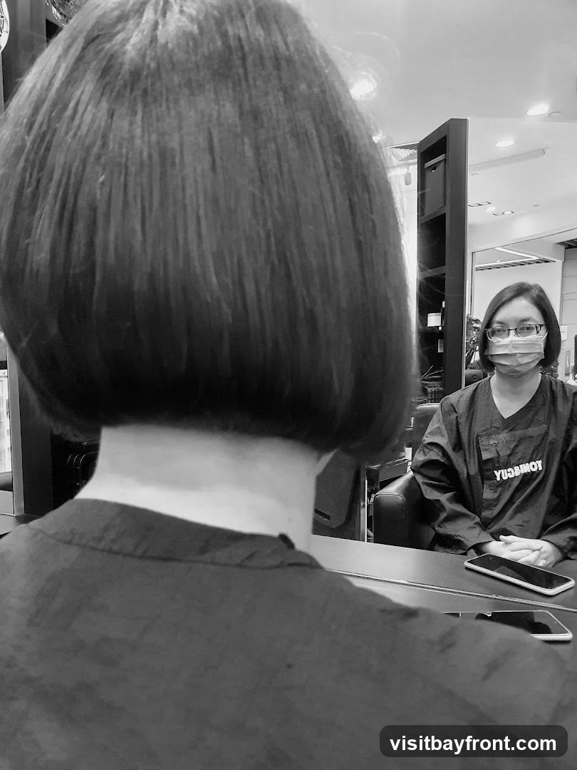 A black and white photo shows the back of a person's head with a bob haircut, reflected in a mirror. In the reflection, a person wearing glasses and a face mask is seated, wearing a dark smock with the word "YUSUMOT" visible on the back. The salon interior is visible in the background, with shelves and chairs.