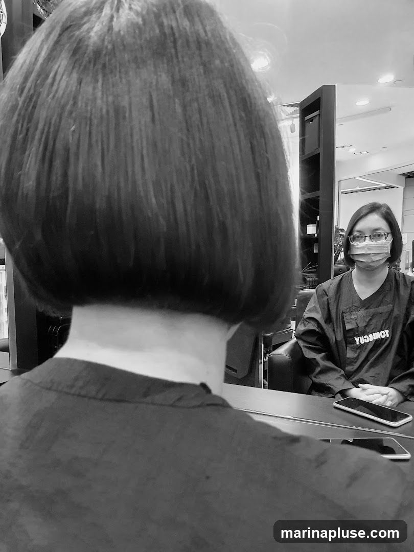 A black and white photo shows the back of a person's head with a bob haircut, reflected in a mirror. In the reflection, a person wearing glasses and a face mask is seated, wearing a dark smock with the word "YUSUMOT" visible on the back. The salon interior is visible in the background, with shelves and chairs.
