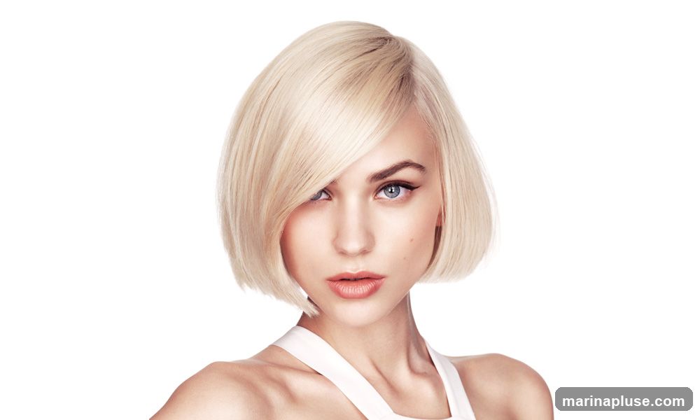 A studio portrait of a young woman with a short, blonde bob hairstyle. She has blue eyes, winged eyeliner, and light pink lipstick. Her hair is styled with a side part and swept over to one side. She is wearing a white top. The background is plain white.